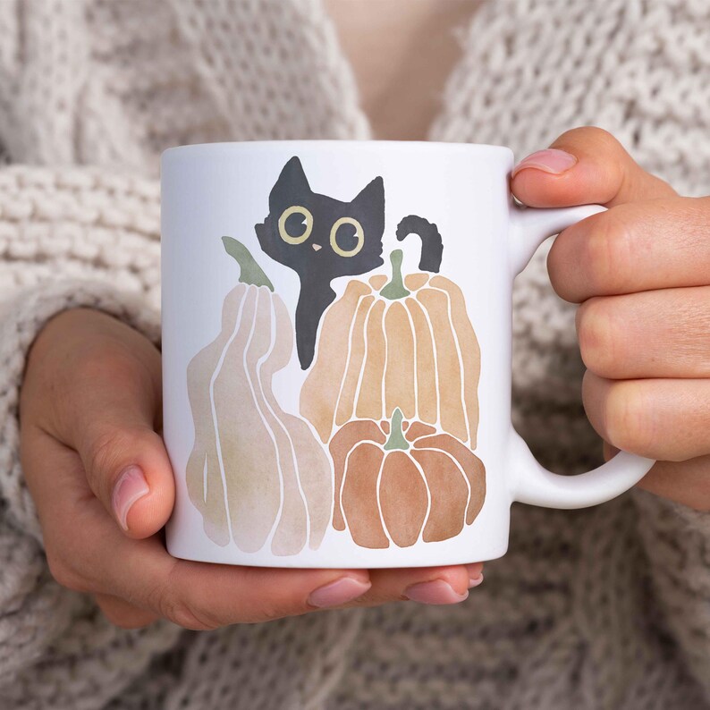 Puede incluir: Taza de cer&aacute;mica blanca con un gato negro asom&aacute;ndose detr&aacute;s de tres calabazas. Las calabazas son naranja, marr&oacute;n y beige con un dise&ntilde;o de acuarela.