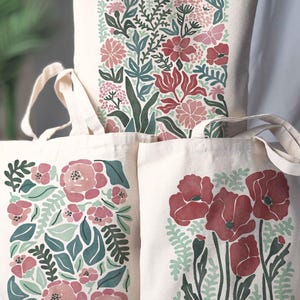 A whimsical floral tote bag with botanical designs in soft pink, light green, and pops of red. One features delicate pink roses with lush greenery, another showcases vibrant red poppies, and the third highlights a spring floral mix with fluttering moths. Perfect for flower lovers, gardeners, and anyone who loves nature-inspired accessories