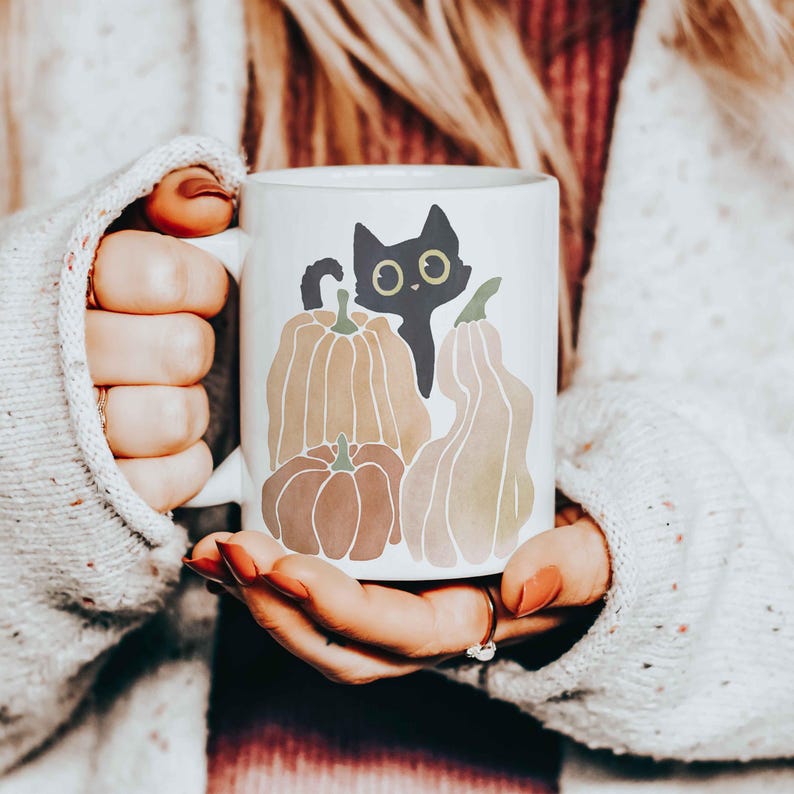 Puede incluir: Taza de cer&aacute;mica blanca con un gato negro asom&aacute;ndose detr&aacute;s de tres calabazas de acuarela. Las calabazas son de color naranja, marr&oacute;n y amarillo.