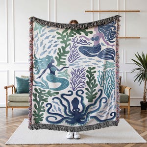 K&ouml;nnte beinhalten: Eine gewebte Wolldecke mit wei&szlig;em Hintergrund und einem bunten Muster aus Meerjungfrauen, einem Oktopus und Seegras. Die Meerjungfrauen sind blau und gr&uuml;n, der Oktopus ist blau und das Seegras ist gr&uuml;n und lila.