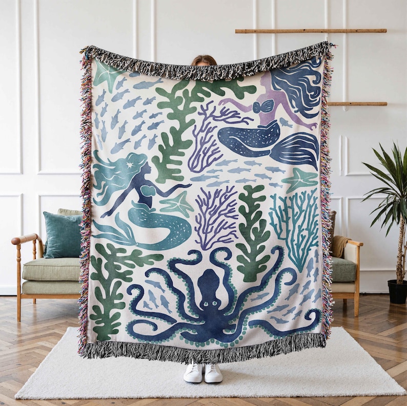 K&ouml;nnte beinhalten: Eine gewebte Wolldecke mit wei&szlig;em Hintergrund und einem bunten Muster aus Meerjungfrauen, einem Oktopus und Seegras. Die Meerjungfrauen sind blau und gr&uuml;n, der Oktopus ist blau und das Seegras ist gr&uuml;n und lila.