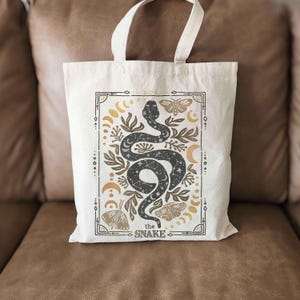 Puede incluir: Una bolsa de tela blanca con un dise&ntilde;o de serpiente negra y el texto "the snake" en un marco dorado. La serpiente est&aacute; rodeada de lunas, estrellas y mariposas.