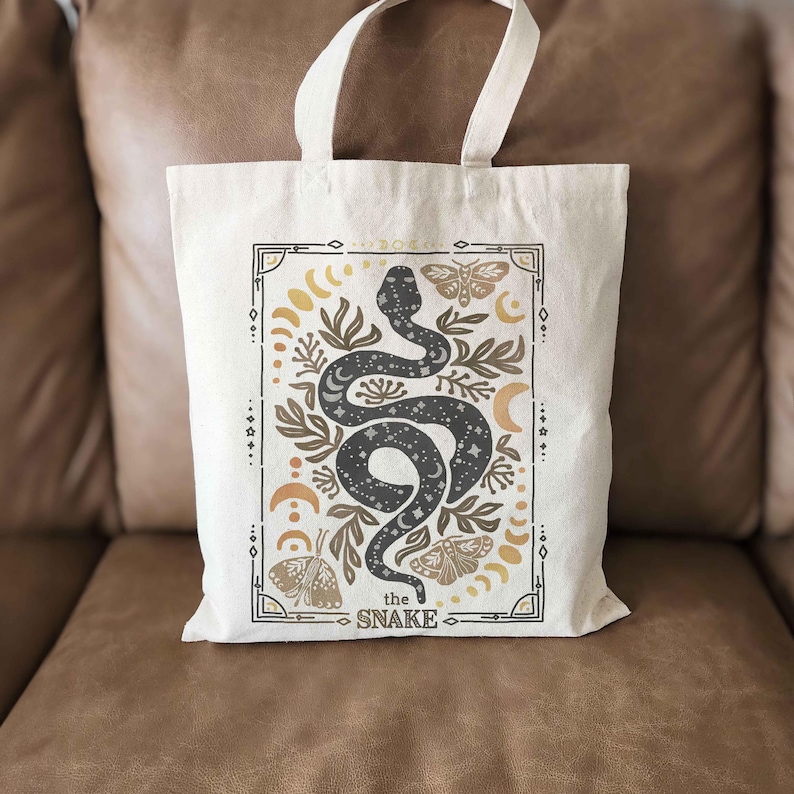 Puede incluir: Una bolsa de tela blanca con un dise&ntilde;o de serpiente negra y el texto "the snake" en un marco dorado. La serpiente est&aacute; rodeada de lunas, estrellas y mariposas.