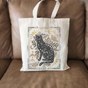 Puede incluir: Una bolsa de tela blanca con una ilustraci&oacute;n de un gato negro. El gato est&aacute; rodeado de lunas, estrellas y flores. El texto "the cat" est&aacute; impreso debajo del gato.