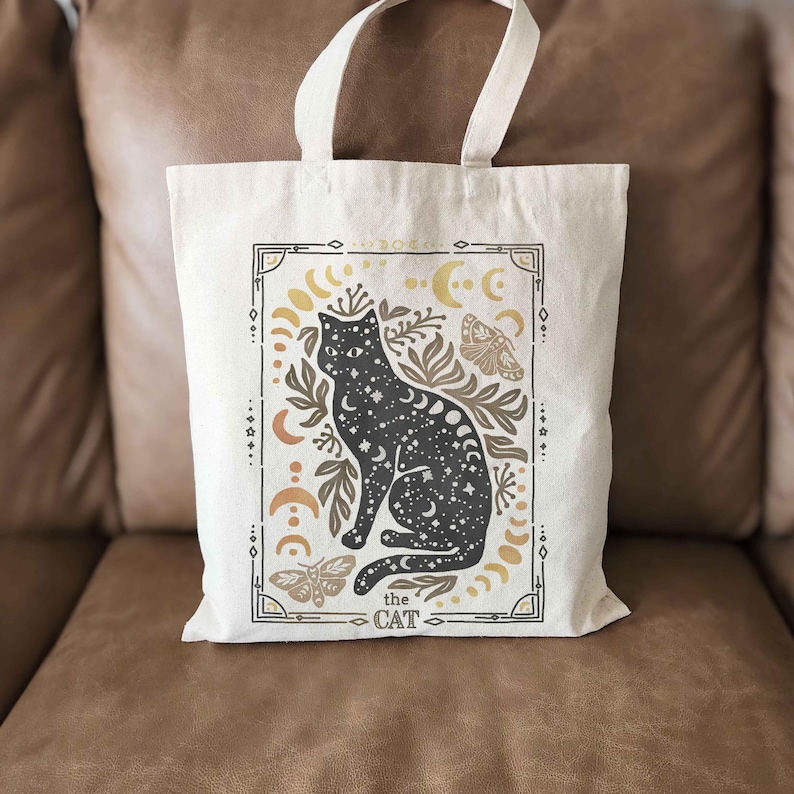 Puede incluir: Una bolsa de tela blanca con una ilustraci&oacute;n de un gato negro. El gato est&aacute; rodeado de lunas, estrellas y flores. El texto "the cat" est&aacute; impreso debajo del gato.