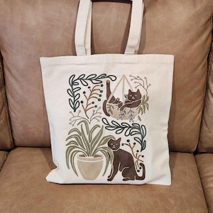 Puede incluir: Una bolsa de lona blanca con un dise&ntilde;o que presenta dos gatos negros, uno en una hamaca colgante y el otro de pie en el suelo, rodeado de follaje verde y bayas marrones.