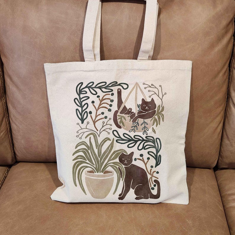 Puede incluir: Una bolsa de lona blanca con un dise&ntilde;o que presenta dos gatos negros, uno en una hamaca colgante y el otro de pie en el suelo, rodeado de follaje verde y bayas marrones.