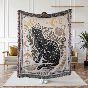 K&ouml;nnte beinhalten: Eine gewebte Wanddecke mit einem schwarzen Katzenmotiv. Die Katze ist von goldenen Monden, Sternen und floralen Elementen umgeben. Der Text "the cat" ist unten auf der Decke aufgedruckt.