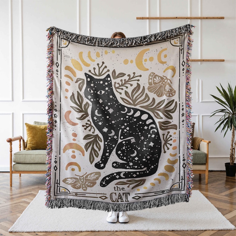 K&ouml;nnte beinhalten: Eine gewebte Wanddecke mit einem schwarzen Katzenmotiv. Die Katze ist von goldenen Monden, Sternen und floralen Elementen umgeben. Der Text "the cat" ist unten auf der Decke aufgedruckt.