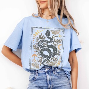 May include: A light blue t-shirt with a graphic print of a snake with stars and moons inside its body. The snake is surrounded by leaves, butterflies, and crescent moons. The graphic is framed with a geometric border.