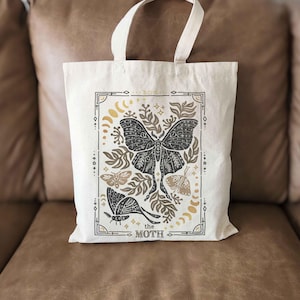 Puede incluir: Una bolsa de tela blanca con un dise&ntilde;o negro y dorado que presenta una polilla, hojas y estrellas. El texto "the moth" est&aacute; impreso en la parte inferior del dise&ntilde;o.