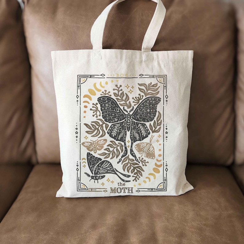 Puede incluir: Una bolsa de tela blanca con un dise&ntilde;o negro y dorado que presenta una polilla, hojas y estrellas. El texto "the moth" est&aacute; impreso en la parte inferior del dise&ntilde;o.