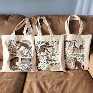 Puede incluir: Tres bolsas de lona marrones con asas negras. Cada bolsa presenta un dise&ntilde;o diferente con gatos marrones, plantas y tazas de caf&eacute;. Los dise&ntilde;os est&aacute;n impresos en marr&oacute;n, verde y blanco.