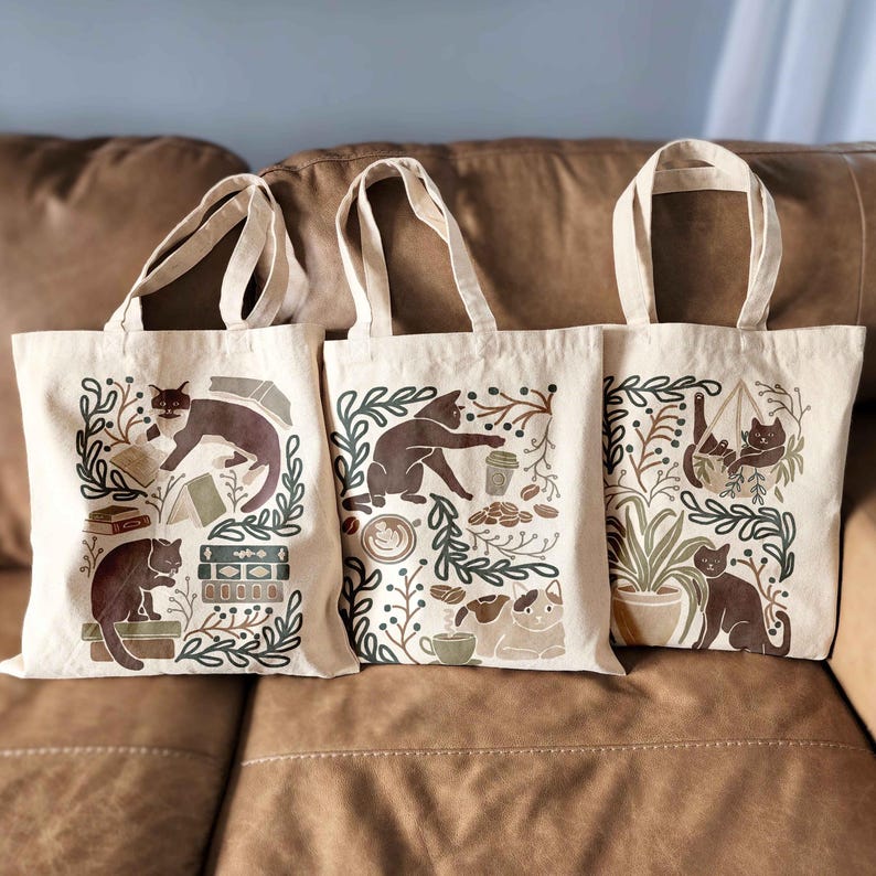 Puede incluir: Tres bolsas de lona marrones con asas negras. Cada bolsa presenta un dise&ntilde;o diferente con gatos marrones, plantas y tazas de caf&eacute;. Los dise&ntilde;os est&aacute;n impresos en marr&oacute;n, verde y blanco.