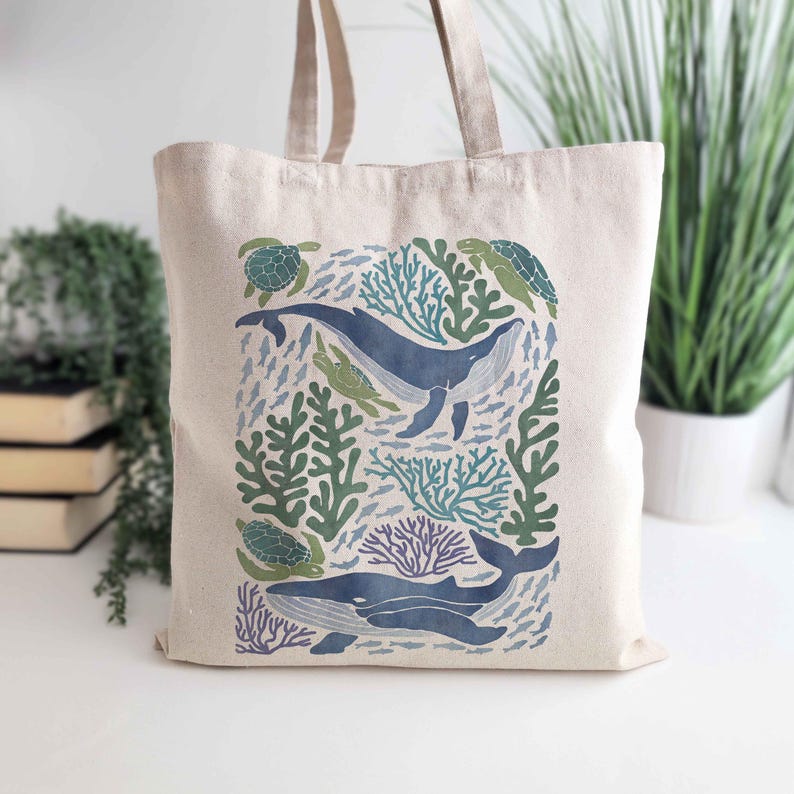 May include: A beige canvas tote bag with a colorful design of whales, sea turtles, and coral reefs. The design is in shades of blue, green, and purple.