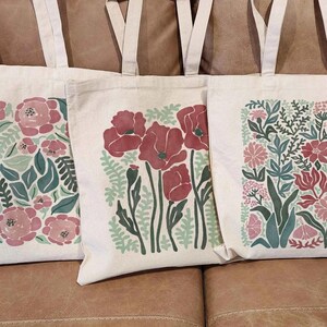 K&ouml;nnte beinhalten: Drei Tragetaschen aus Canvas mit floralen Designs. Die Taschen haben cremefarbene Griffe und Hintergr&uuml;nde. Die floralen Muster umfassen rosa und rote Blumen mit gr&uuml;nen Bl&auml;ttern. Die Taschen liegen auf einem braunen Ledersofa.