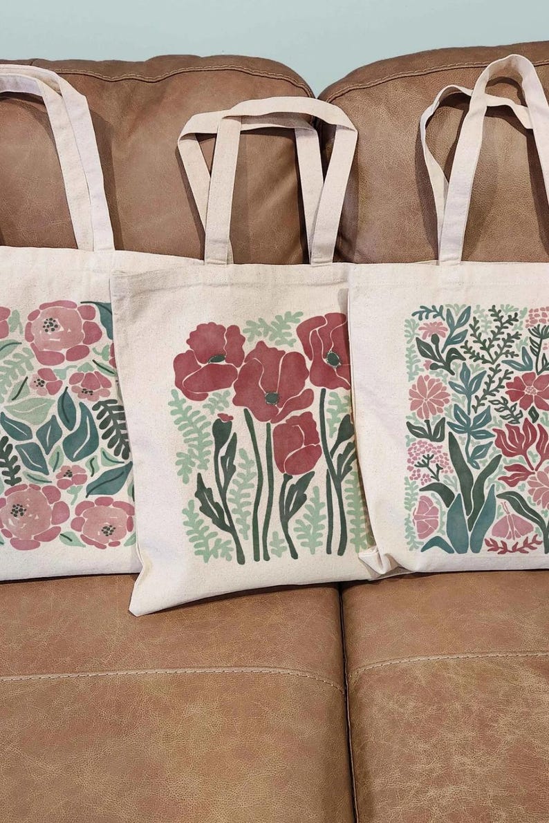 K&ouml;nnte beinhalten: Drei Tragetaschen aus Canvas mit floralen Designs. Die Taschen haben cremefarbene Griffe und Hintergr&uuml;nde. Die floralen Muster umfassen rosa und rote Blumen mit gr&uuml;nen Bl&auml;ttern. Die Taschen liegen auf einem braunen Ledersofa.