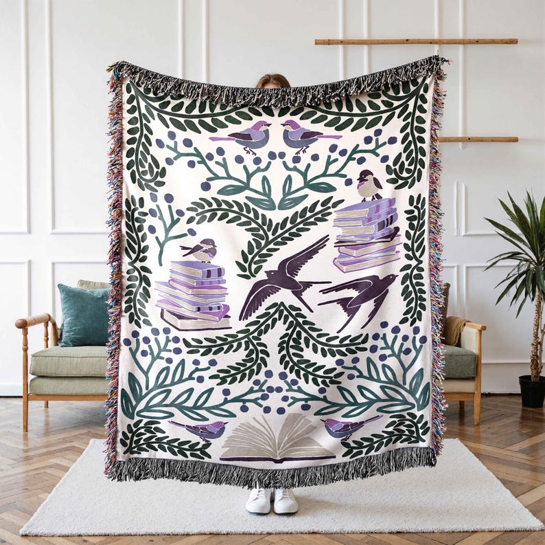 May include: A white woven throw blanket with a green and purple floral pattern featuring birds and stacks of books. The blanket has a fringe border.