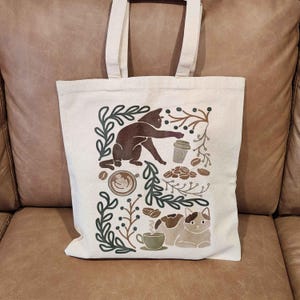 Puede incluir: Una bolsa de tela blanca con un gato marr&oacute;n, granos de caf&eacute; y una taza de caf&eacute; impresos en ella. El dise&ntilde;o est&aacute; rodeado de hojas y ramas verdes.