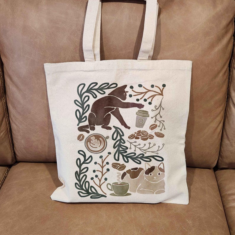 Puede incluir: Una bolsa de tela blanca con un gato marr&oacute;n, granos de caf&eacute; y una taza de caf&eacute; impresos en ella. El dise&ntilde;o est&aacute; rodeado de hojas y ramas verdes.
