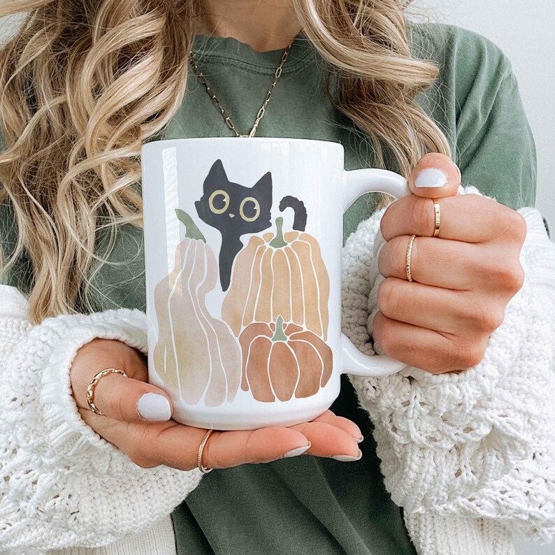 Puede incluir: Taza de cer&aacute;mica blanca con un gato negro asom&aacute;ndose detr&aacute;s de tres calabazas. Las calabazas son naranja, marr&oacute;n y beige. El gato tiene ojos verdes.