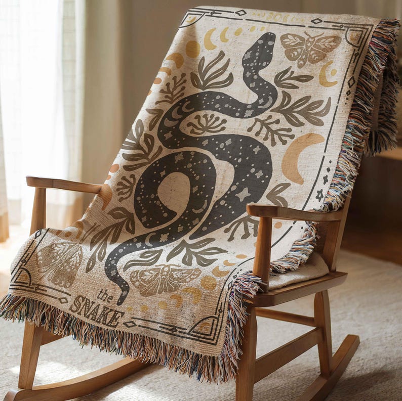 Puede incluir: Una manta tejida con un dise&ntilde;o de serpiente negra, rodeada de lunas, estrellas y polillas. El texto "the snake" est&aacute; impreso en la manta.
