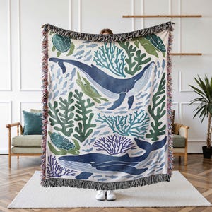 K&ouml;nnte beinhalten: Eine gewebte Wolldecke mit einem Muster aus einem blauen Wal, Meeresschildkr&ouml;ten, Korallen und Algen auf wei&szlig;em Hintergrund. Die Decke hat einen Fransenrand.