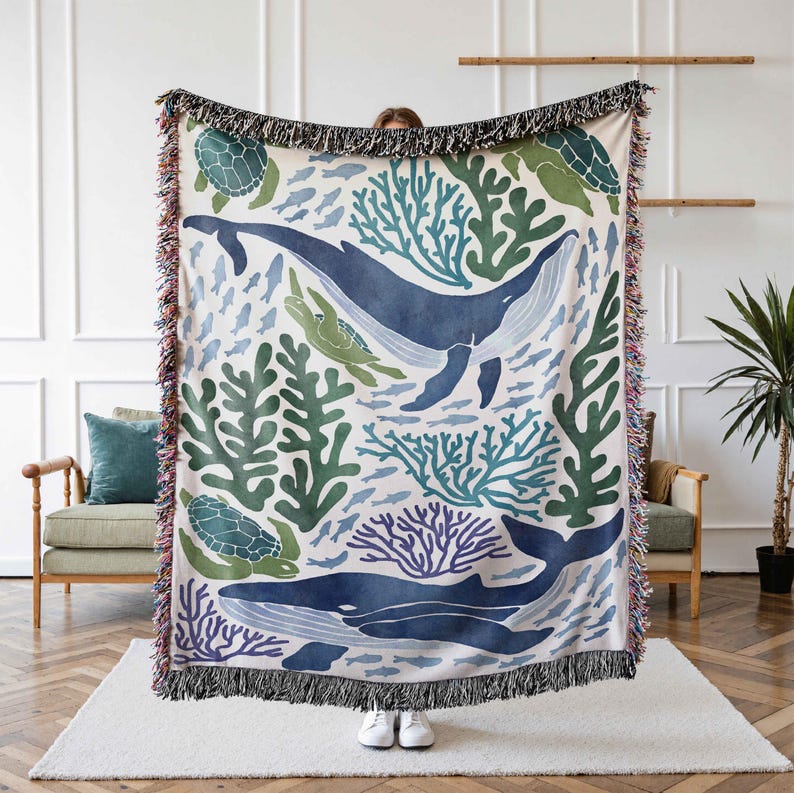 K&ouml;nnte beinhalten: Eine gewebte Wolldecke mit einem Muster aus einem blauen Wal, Meeresschildkr&ouml;ten, Korallen und Algen auf wei&szlig;em Hintergrund. Die Decke hat einen Fransenrand.
