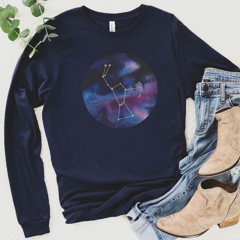 K&ouml;nnte beinhalten: Ein marineblaues Langarmshirt mit einem Aquarell-Galaxie-Druck, der eine Konstellation in Gold zeigt. Das Shirt liegt auf einer wei&szlig;en Oberfl&auml;che mit einem Paar braunen Stiefeletten und einer blauen Jeans.