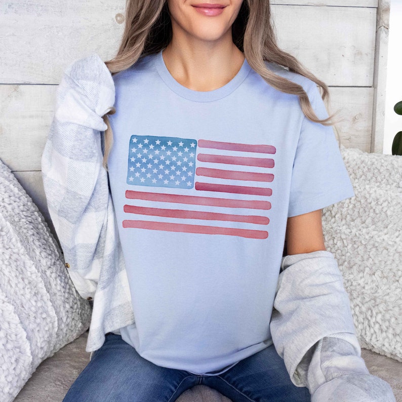 May include: A light blue t-shirt with a watercolor design of the American flag. The flag is red, white, and blue with a faded, watercolor effect.