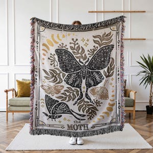 K&ouml;nnte beinhalten: Eine gewebte Tapisserie-Decke mit einem schwarzen und wei&szlig;en Motten-Design. Die Motte ist von goldenem und braunem Laub umgeben und der Text "the moth" ist unten aufgedruckt.