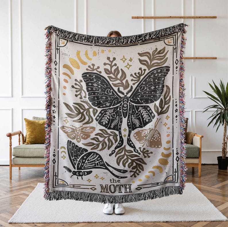 K&ouml;nnte beinhalten: Eine gewebte Tapisserie-Decke mit einem schwarzen und wei&szlig;en Motten-Design. Die Motte ist von goldenem und braunem Laub umgeben und der Text "the moth" ist unten aufgedruckt.