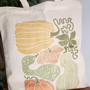Puede incluir: Bolso tote de color crema con una colorida ilustraci&oacute;n de calabazas y calabacines en tonos amarillos, naranjas y verdes. El bolso tiene asas resistentes y est&aacute; hecho de tela duradera. Ideal para ir de compras.