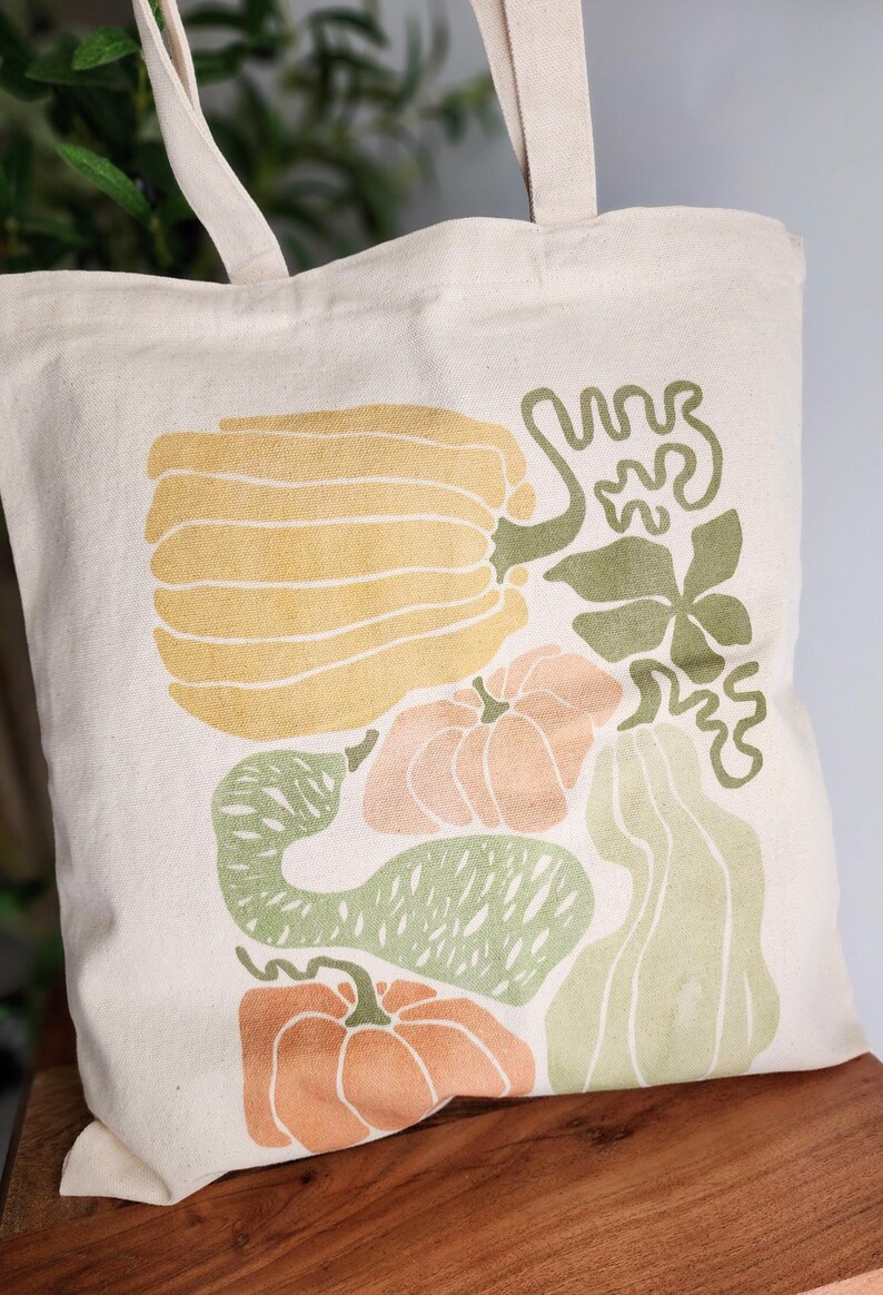 Puede incluir: Bolso tote de color crema con una colorida ilustraci&oacute;n de calabazas y calabacines en tonos amarillos, naranjas y verdes. El bolso tiene asas resistentes y est&aacute; hecho de tela duradera. Ideal para ir de compras.
