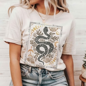 May include: A white t-shirt with a graphic print of a snake with stars and moons. The graphic is surrounded by a border with the text "the snake".