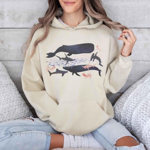 K&ouml;nnte beinhalten: Ein cremefarbener Hoodie mit einem grafischen Druck eines Wals, Hais und Quallen in Blaut&ouml;nen, Rosa und Grau.