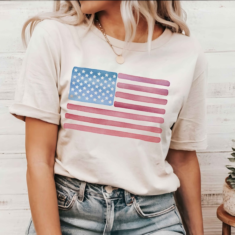 May include: A white t-shirt with a watercolor American flag design. The flag is in shades of blue, red, and pink.