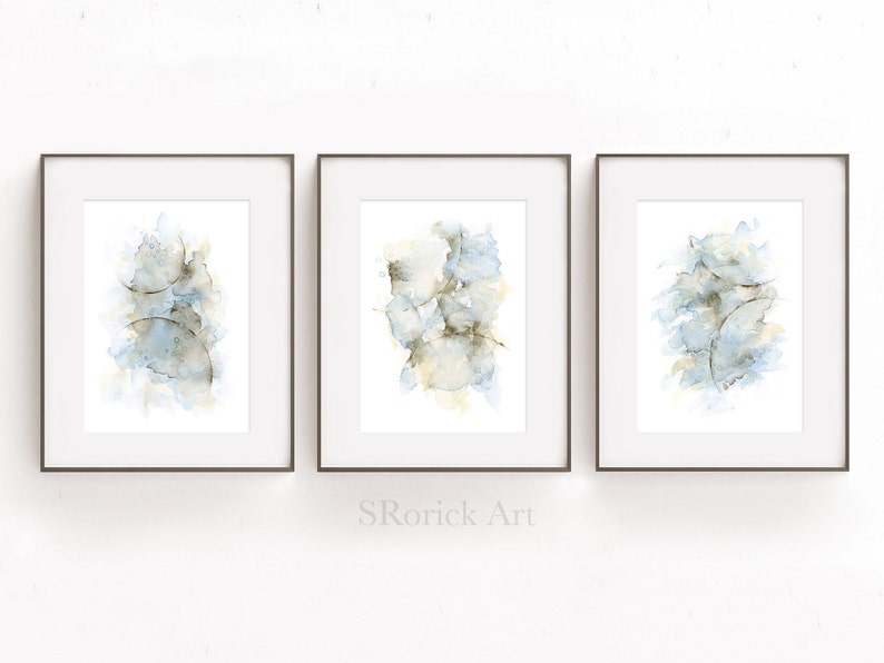 May include: Three framed watercolor paintings with abstract designs in shades of blue and brown. The paintings are displayed on a white wall.