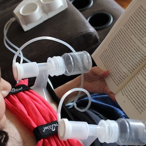 May include: A woman using a double electric breast pump while reading a book. The pump has clear plastic bottles and tubing, with a red fabric support. A white accessory holds additional bottles. The book is open, displaying text.