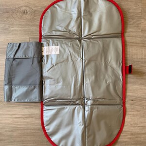 May include: A gray and red baby changing pad, open and unfolded. The pad has a silver interior and a red border. A small, attached gray pouch is on the left side. The pad is designed for on-the-go diaper changes.