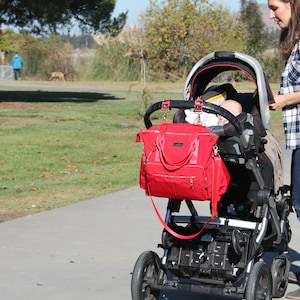 May include: A red baby diaper bag with gold zippers hangs from the handle of a black and gray stroller. The stroller has a black canopy and a baby is inside. A person wearing a plaid shirt and jeans stands next to the stroller.