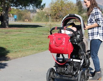 May include: A red baby diaper bag with gold zippers hangs from the handle of a black and gray stroller. The stroller has a black canopy and a baby is inside. A person wearing a plaid shirt and jeans stands next to the stroller.