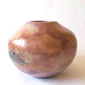 May include: A handcrafted, round ceramic vase with a wide opening. The vase has a glossy finish with a blend of earthy tones, including shades of brown, copper, and hints of gray. The overall shape is smooth and organic.