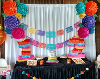 May include: A festive party scene with colorful decorations. Large paper flowers in orange, teal, purple, and pink adorn the backdrop. A table displays a cake, piñatas shaped like numbers, and decorative banners. The text on the banner reads "Quinn is 1" and "Evelyn is 1".