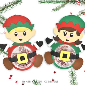 Puede incluir: Dos porta golosinas con temática de duendes navideños, uno con cabello rubio y otro con cabello castaño. Ambos duendes llevan gorros verdes y rojos y tienen un bastón de caramelo en sus manos. Los porta golosinas están hechos de papel y tienen un círculo de plástico transparente en el medio para guardar dulces.