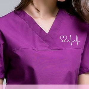 May include: A purple medical scrub top with a white heart and EKG line design. The design is embroidered on the chest of the scrub top.