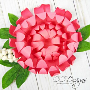 May include: A large, pink paper flower with multiple layers of petals. The flower is surrounded by green leaves.
