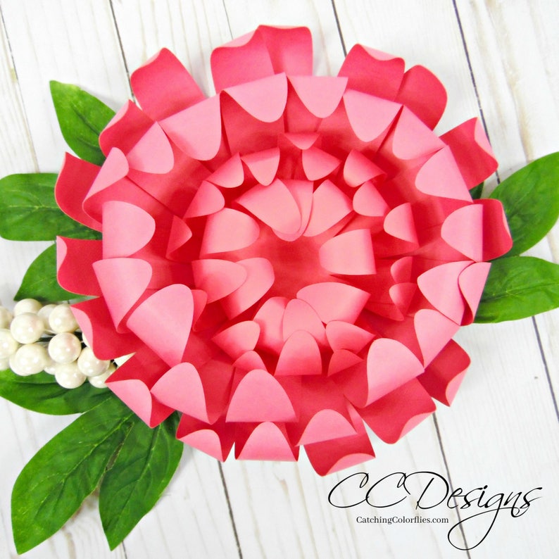 May include: A large, pink paper flower with multiple layers of petals. The flower is surrounded by green leaves.