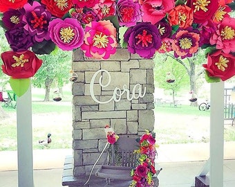 May include: A brick wall decorated with a large arrangement of paper flowers in shades of pink, red, and purple. The flowers are arranged in a cascading pattern and spell out the name "Cora" in white letters. A small wooden chair is decorated with a garland of flowers.