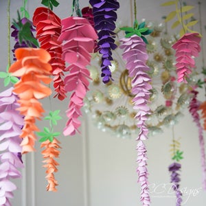 May include: A collection of paper wisteria flowers in various colours, including pink, purple, orange, and red, hanging from the ceiling. The flowers are made of paper and are arranged in clusters.